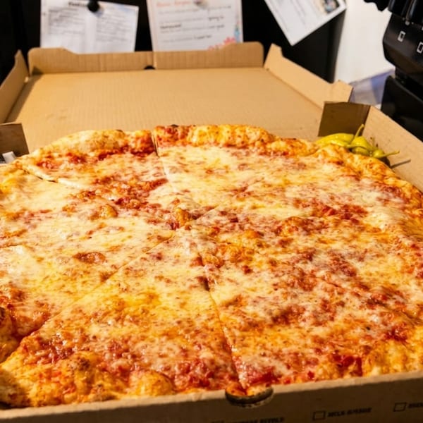 A large cheese pizza in a cardboard box with dipping sauces and peppers, next to a stove.