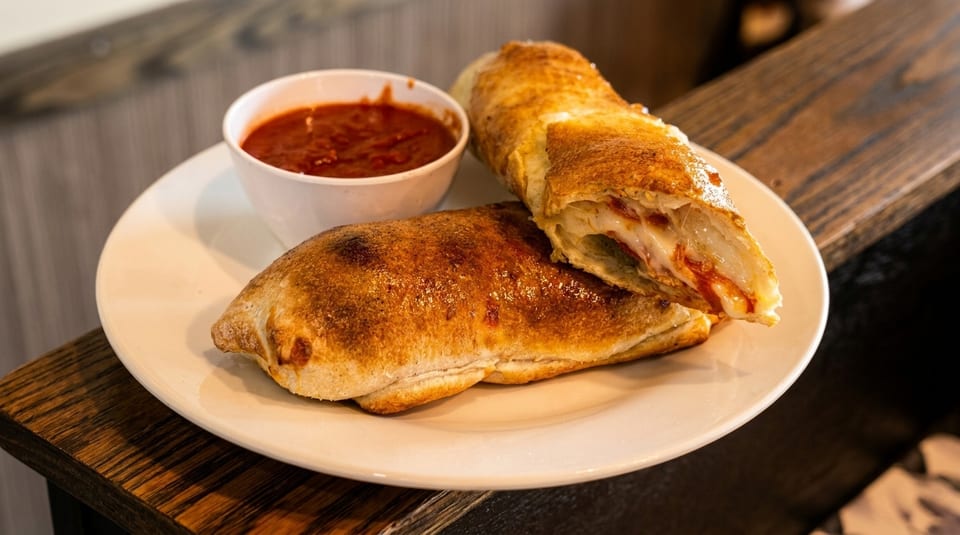 Two golden-brown strombolis, one sliced open revealing melted cheese and sauce, with a side of marinara.