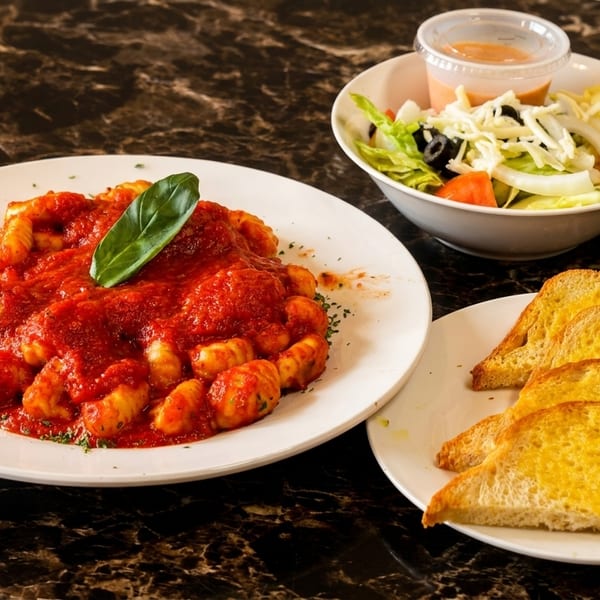 Gnocchi with red sauce and basil, a side salad, and garlic bread on a marble table.